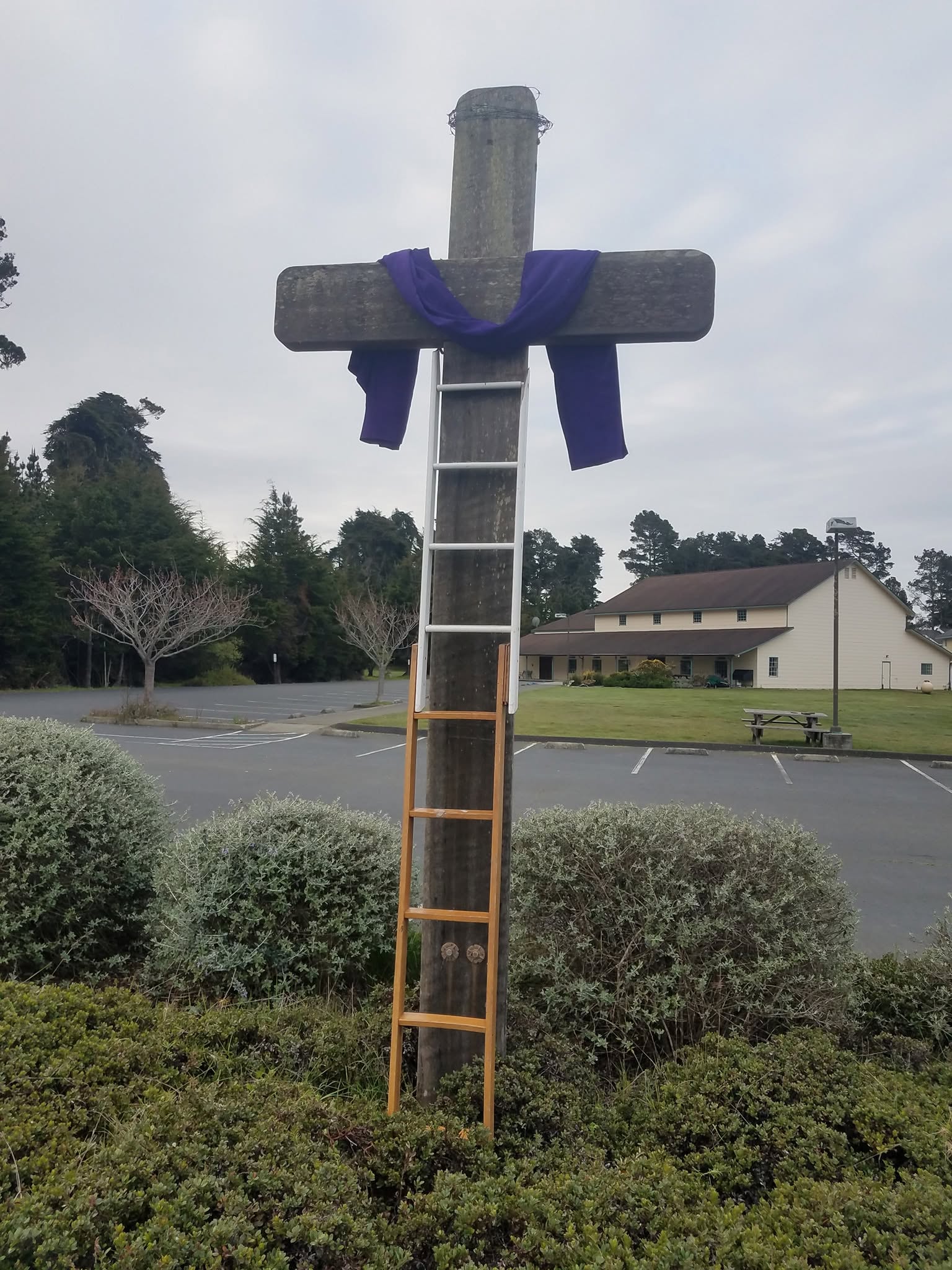Wooden cross on the church grounds with the building and evergreen trees beyond